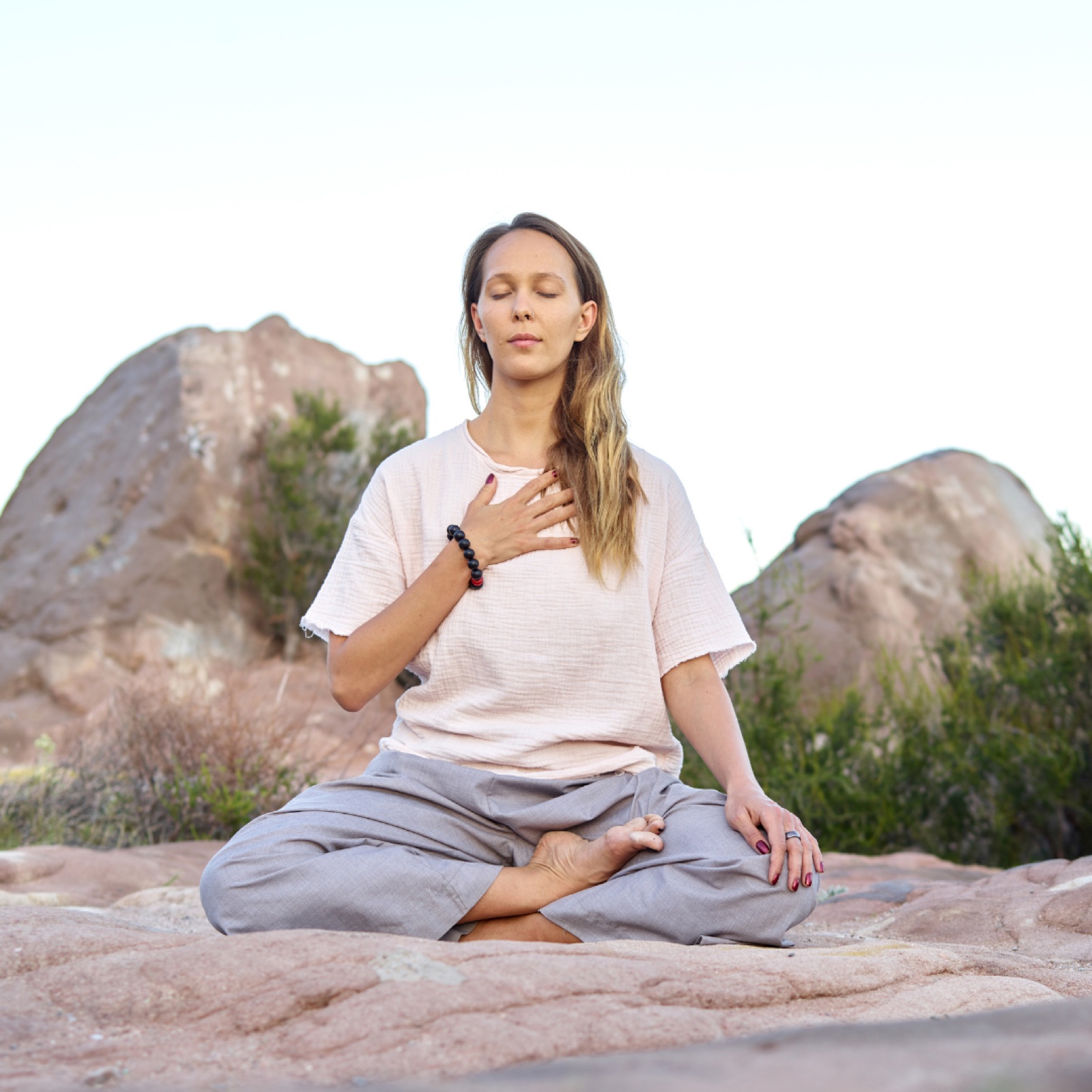 Woman meditating outdoors on rocky terrain, eyes closed, hand on chest. Wearing light clothes, peaceful mood, natural background.