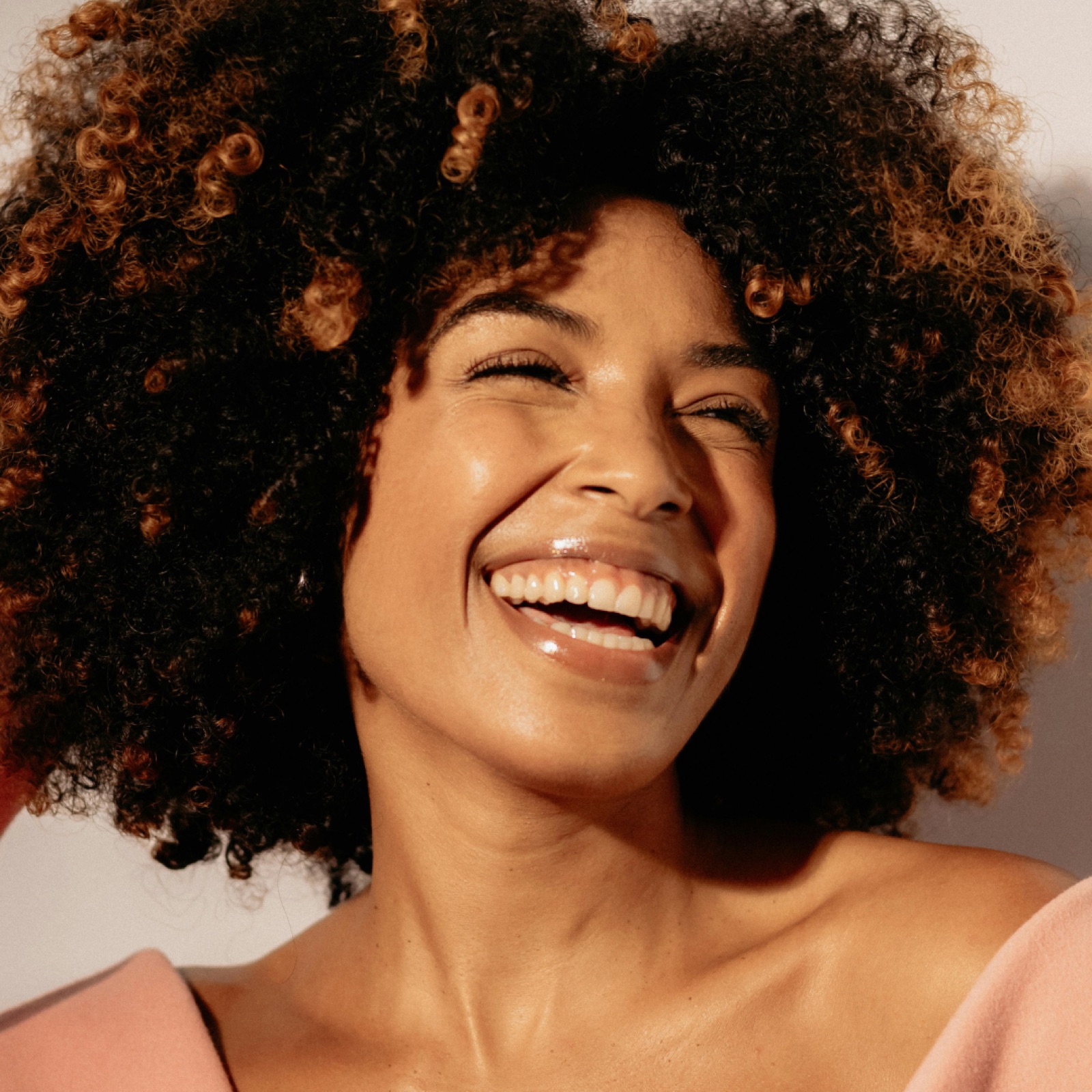 Woman with curly hair laughing, wearing a pink top. Warm lighting creates a joyful mood against a neutral background.