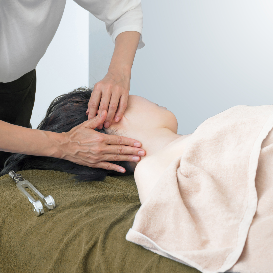 Person receiving a head massage on a brown towel, with a calm setting. Another person's hands gently press near the ear, conveying relaxation. Tuning fork sitting nearby.