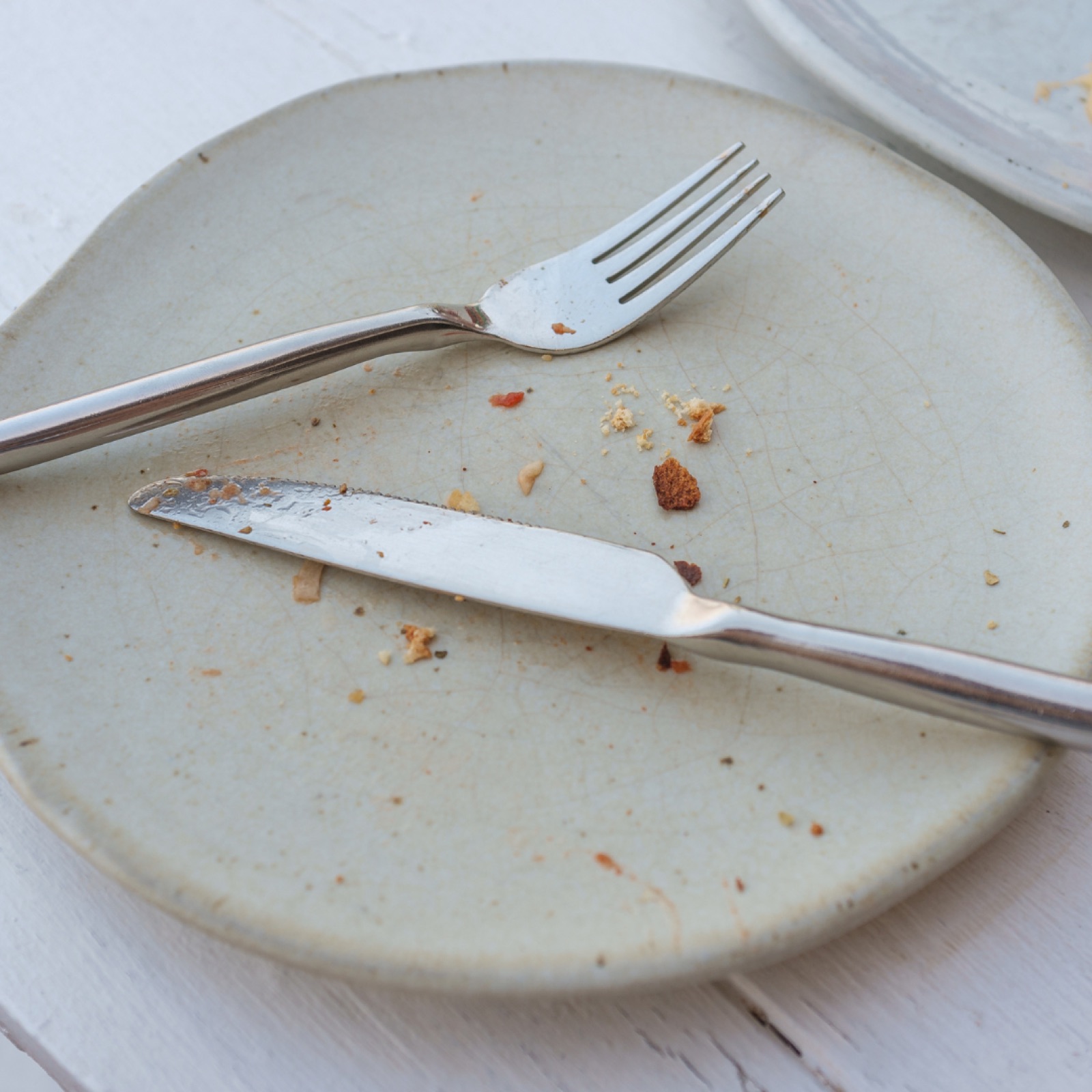 Empty plate with crumbs, a fork, and a knife on a white wooden table. Light gray plate creating a minimal, unfinished meal vibe.