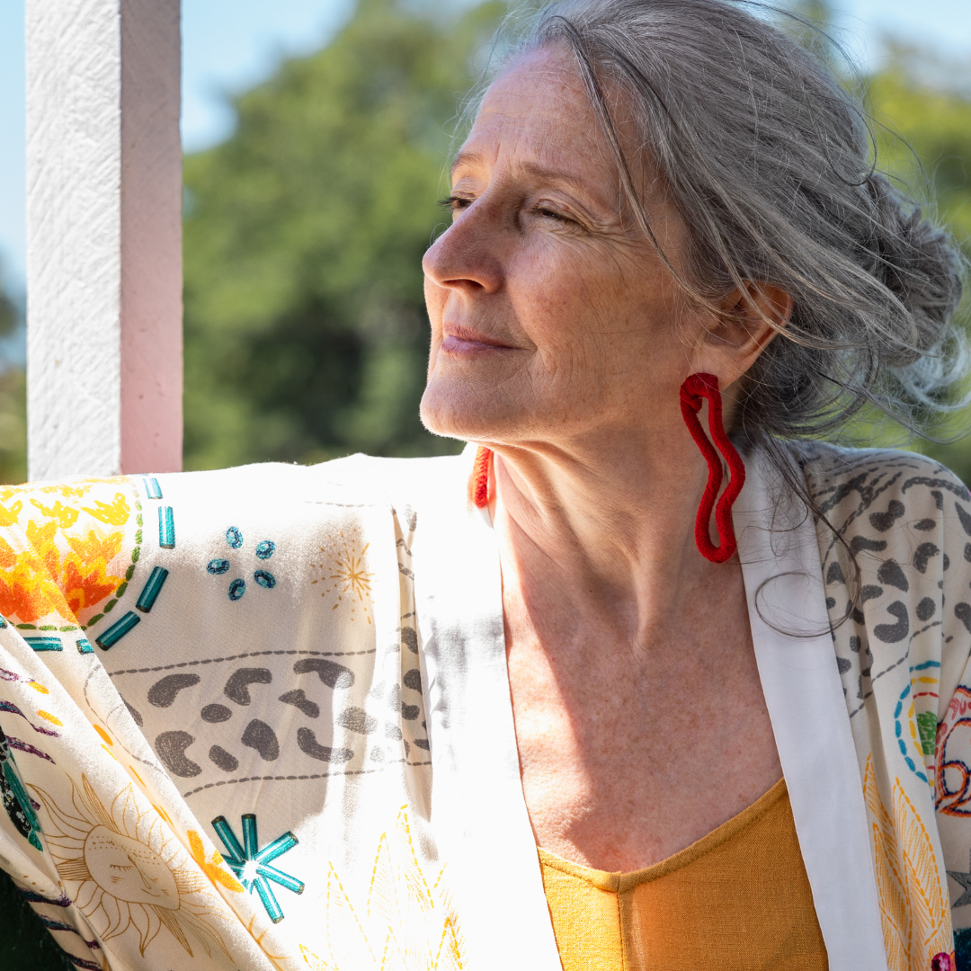 Woman with gray hair looks contently into the distance, wearing a colorful patterned shawl and red earrings, outdoors with greenery.