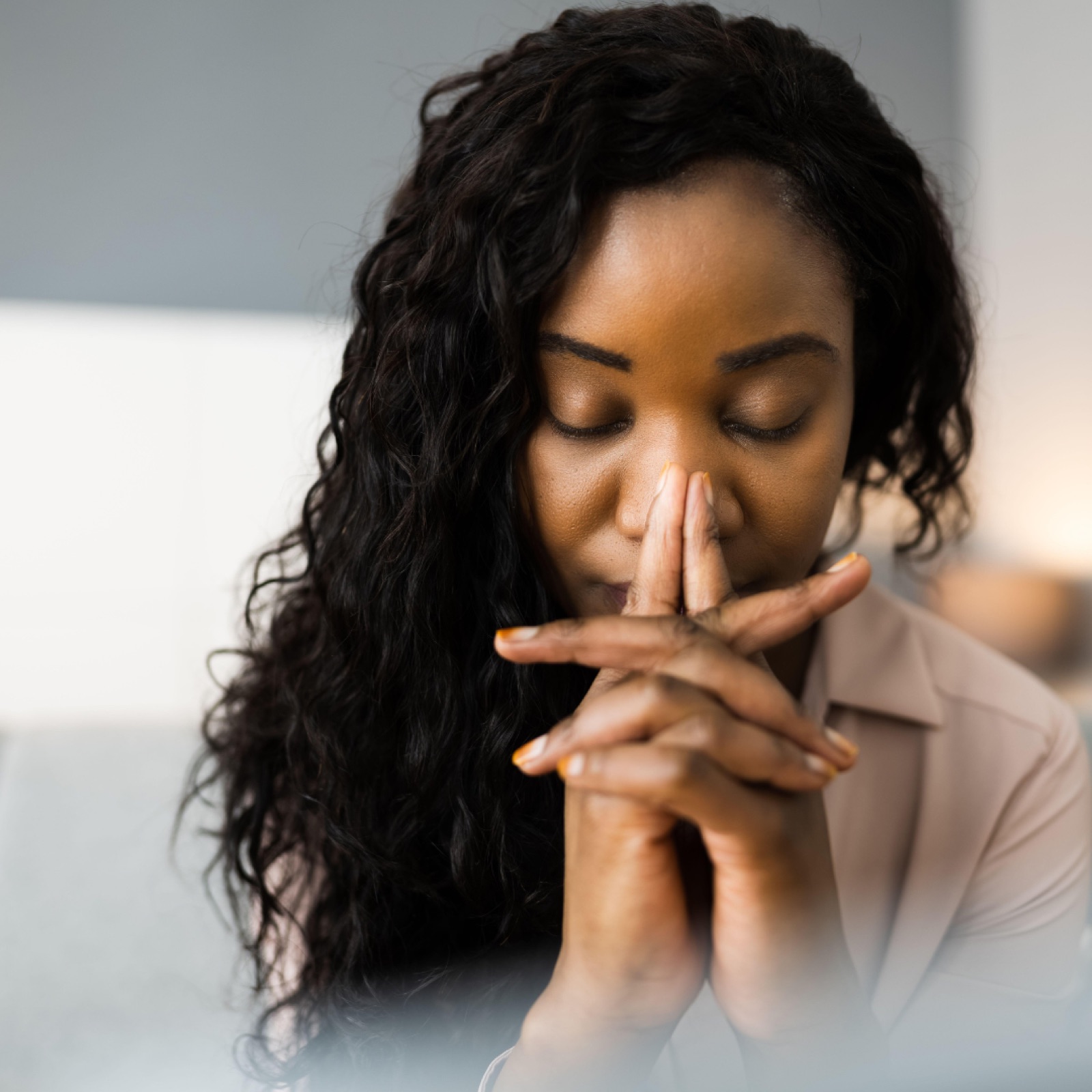 Woman with curly hair, eyes closed, resting face on clasped hands in a contemplative mood, indoors with soft lighting.