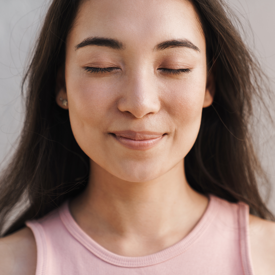 Woman with long hair and closed eyes, smiling peacefully. Wears a pink top against a neutral background. Calm and serene mood.