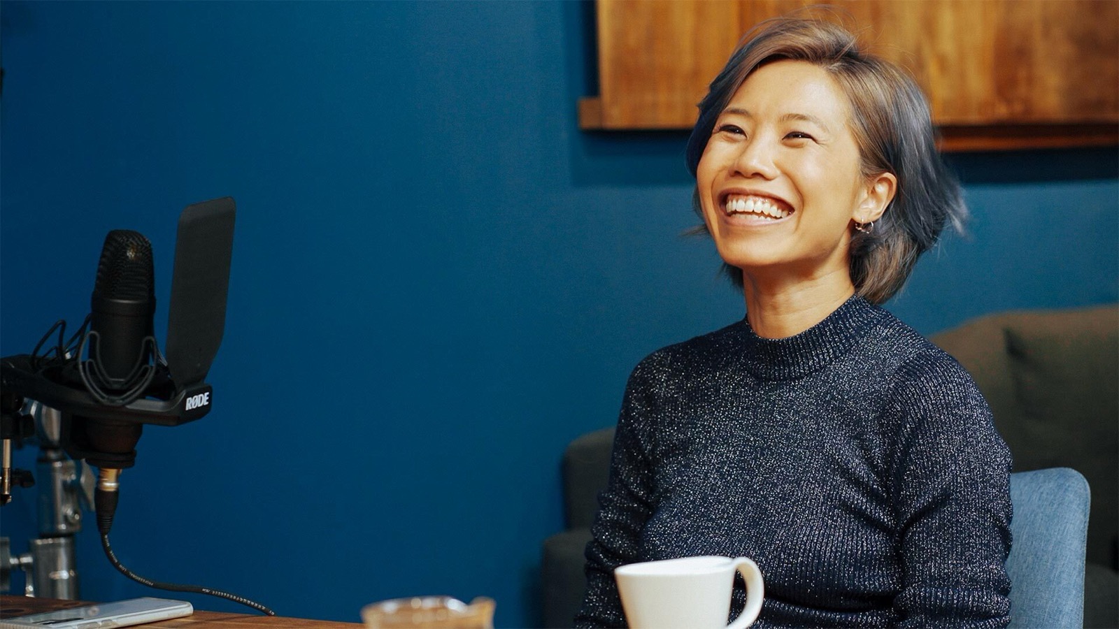 Smiling person with short hair sits at a table with a cup, microphone nearby, in a cozy room with blue walls and wooden decor.