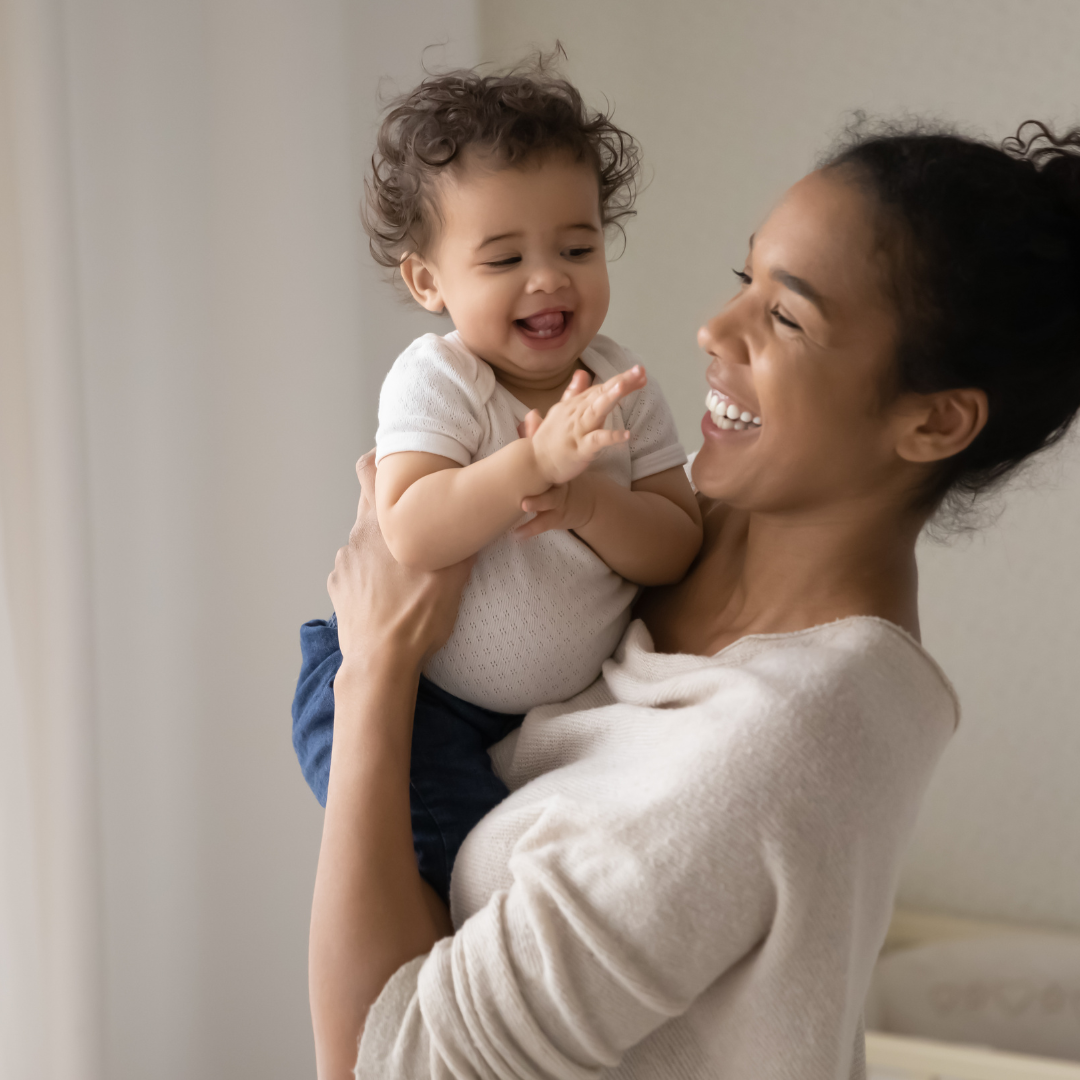 A woman joyfully holds a smiling baby in a softly lit room. Both wear light-colored clothing, creating a warm, happy atmosphere.