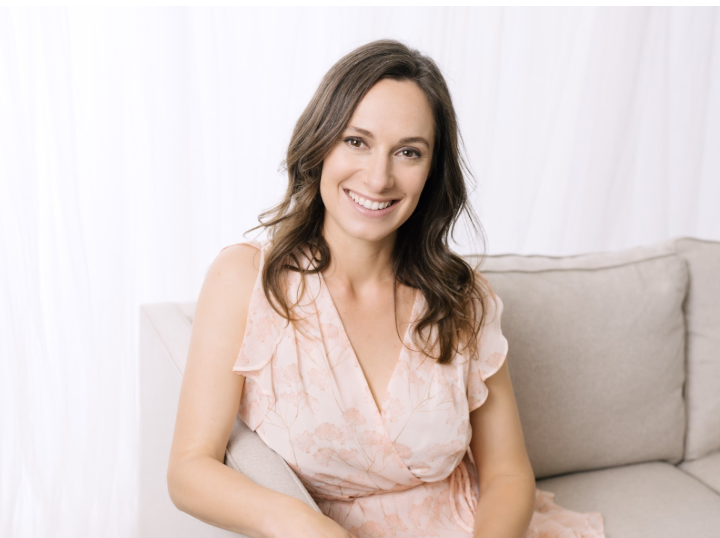 Smiling woman in a floral, light pink dress sits on a beige sofa with a white curtain backdrop, creating a bright and cheerful setting.