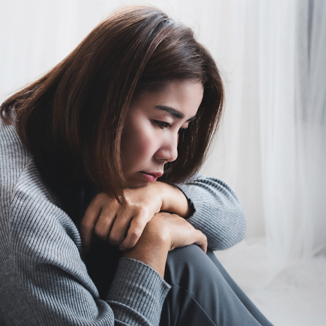 Woman in a gray sweater sits pensively with clasped hands against knees. Soft, light background conveys a contemplative mood.