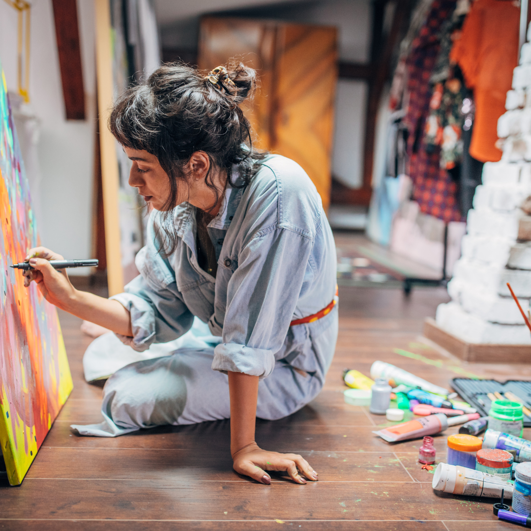 Artist in denim overalls painting a colorful canvas on a wooden floor. Paints scattered nearby. Cozy studio with a creative mood.