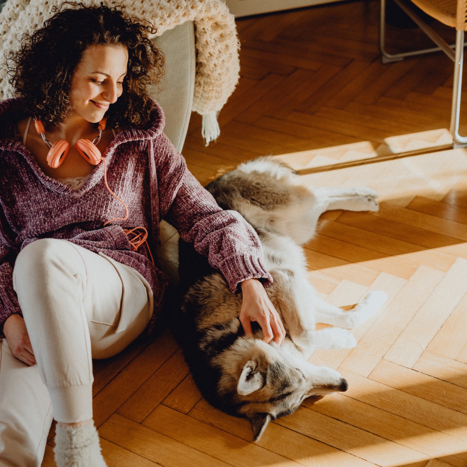 A woman in a purple sweater with headphones smiles while sitting on a wooden floor, petting a relaxed dog in sunlight. Cozy atmosphere.