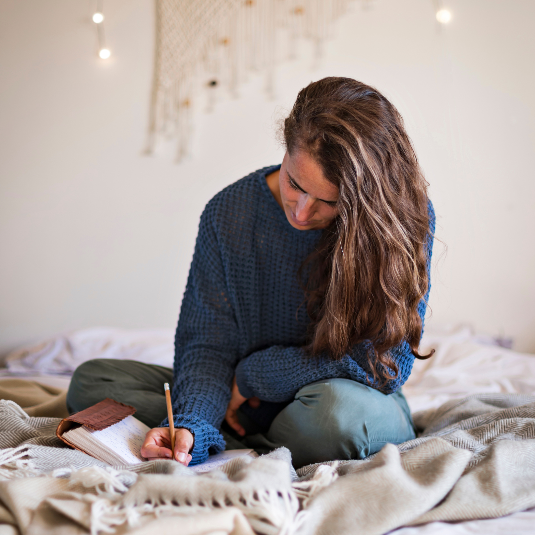 Woman sitting on a bed, writing in a notebook with a pencil. She's wearing a blue sweater. Soft lighting and cozy, relaxed atmosphere.