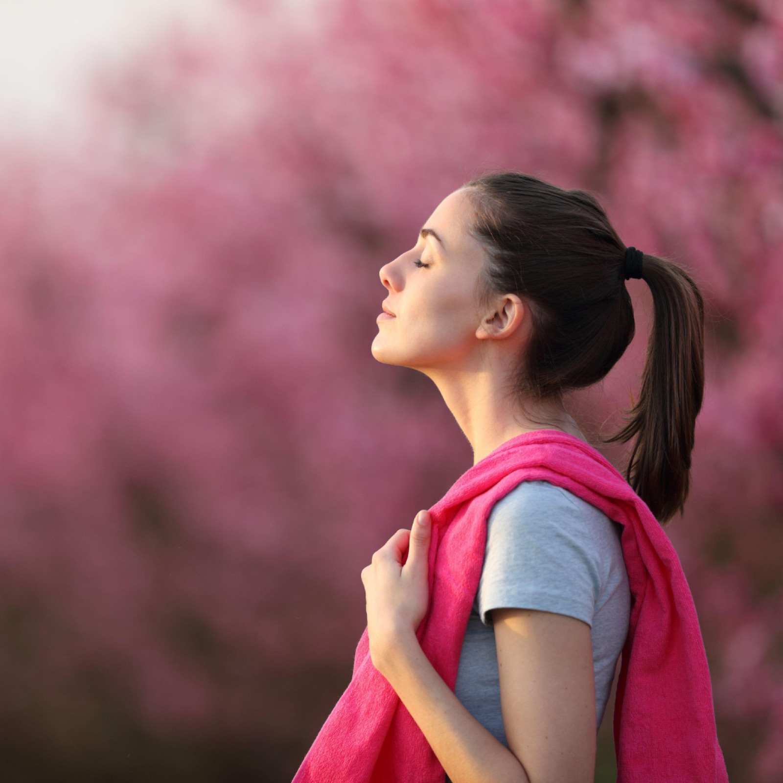 Woman with closed eyes in profile, draped in pink, stands amidst a pink blossom background, exuding calmness in soft sunlight.