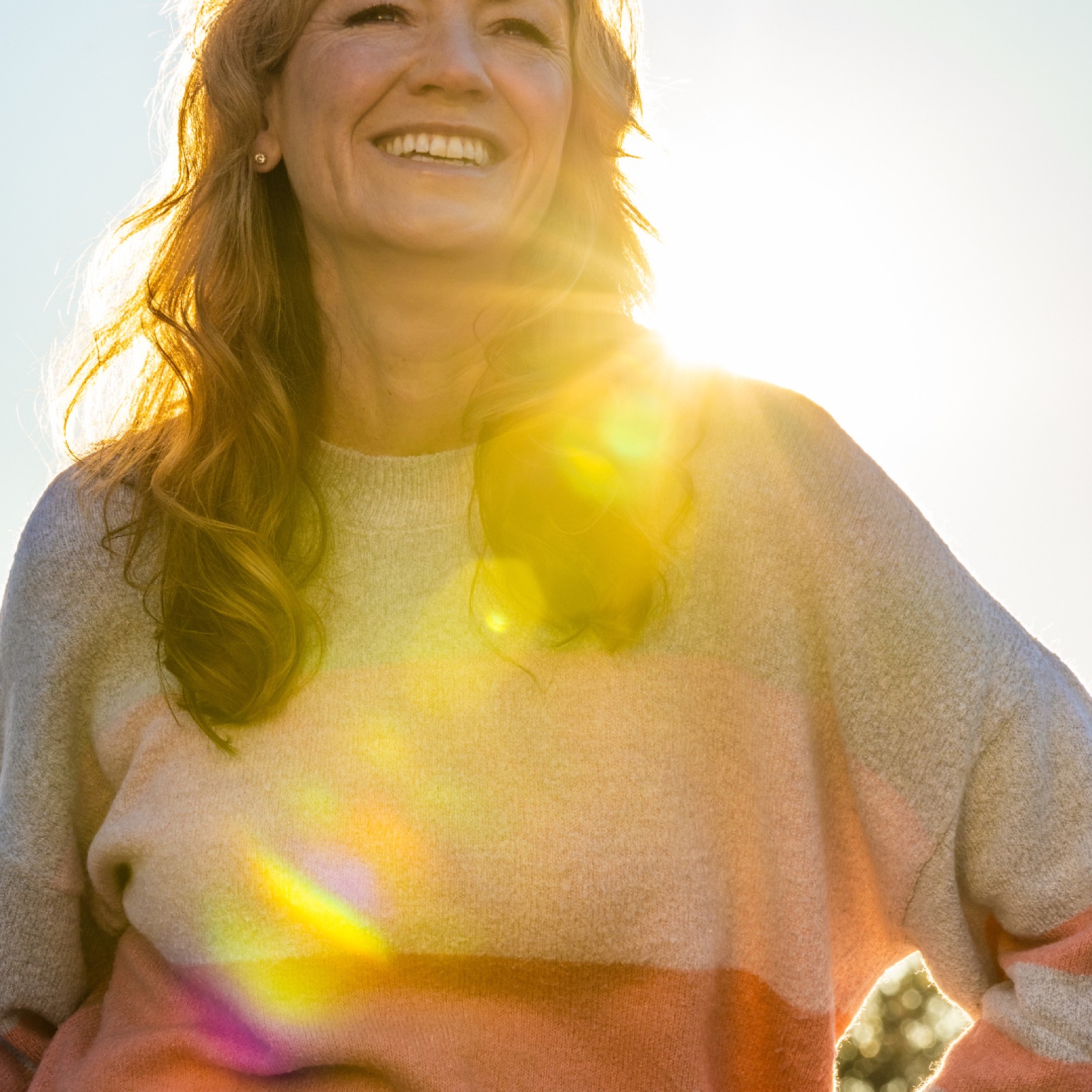 Smiling woman in a colorful sweater stands outdoors, bathed in warm sunlight. Sunlight creates lens flares, adding a cheerful mood.