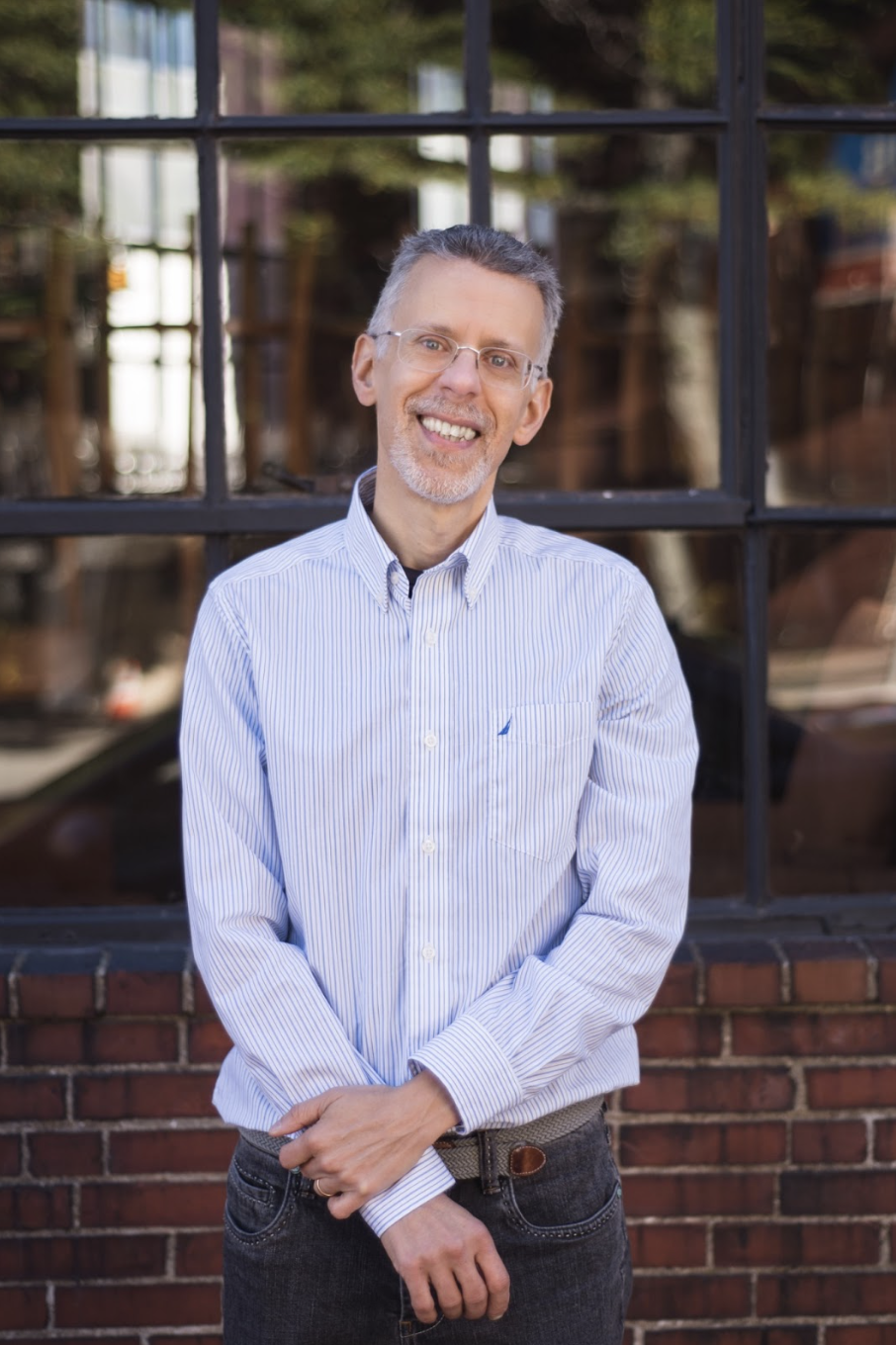 Smiling man in a striped white shirt stands in front of a window with brick trim. Background shows blurred outdoor reflections, creating a warm atmosphere.