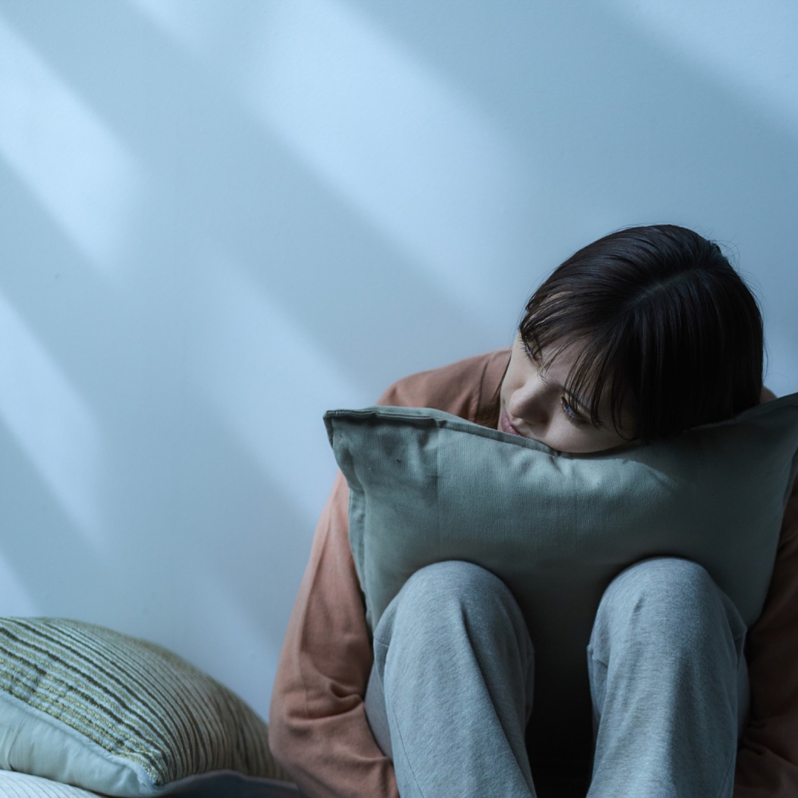 Woman sitting against a blue wall, hugging a pillow, looking contemplative. Soft light casts shadows, creating a calm, introspective mood.