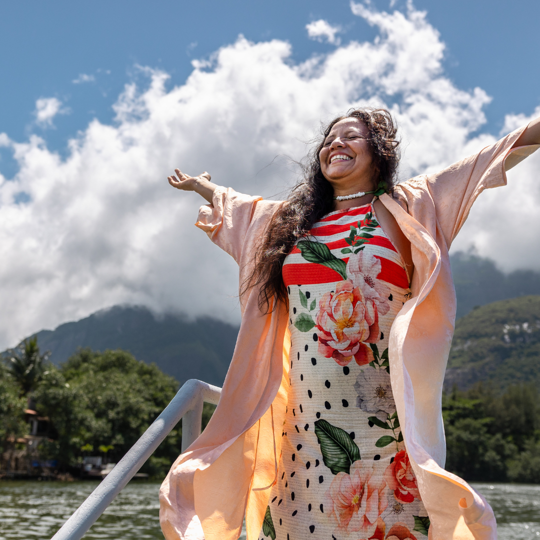 Woman joyfully raises arms on a boat, wearing a floral dress and peach robe. Cloudy mountains and lush greenery in the background.