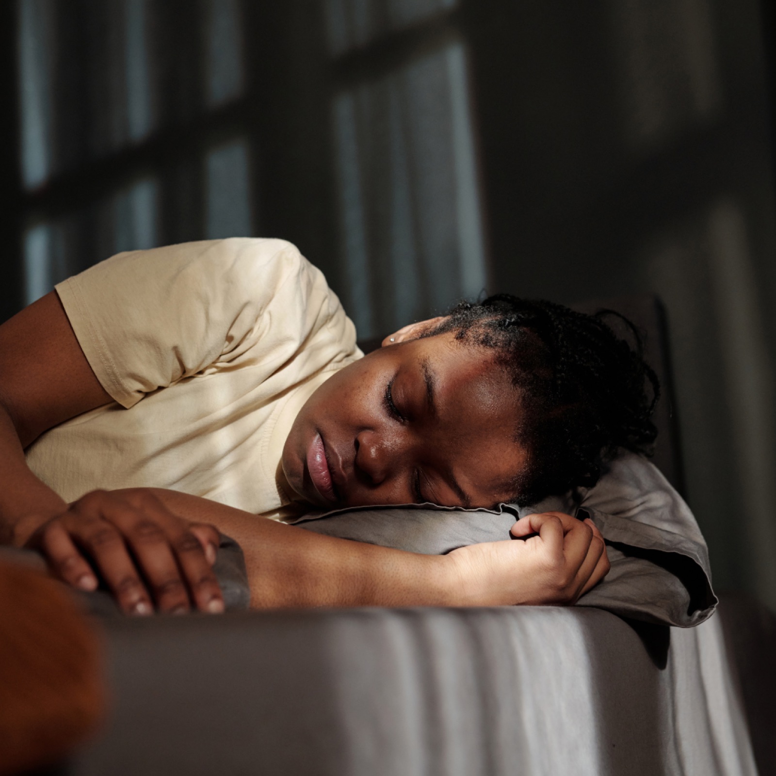 Person sleeping in daylight on a bed with gray sheets, wearing a light-colored shirt. Soft shadows and a calm, peaceful mood.