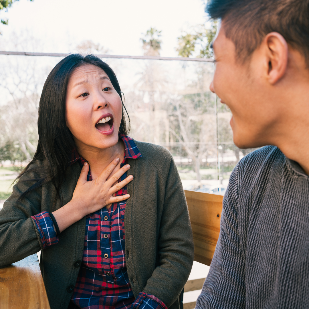 Woman gestures passionately while speaking to a man outdoors. She's wearing a plaid shirt and gray jacket. Trees in the blurred background.