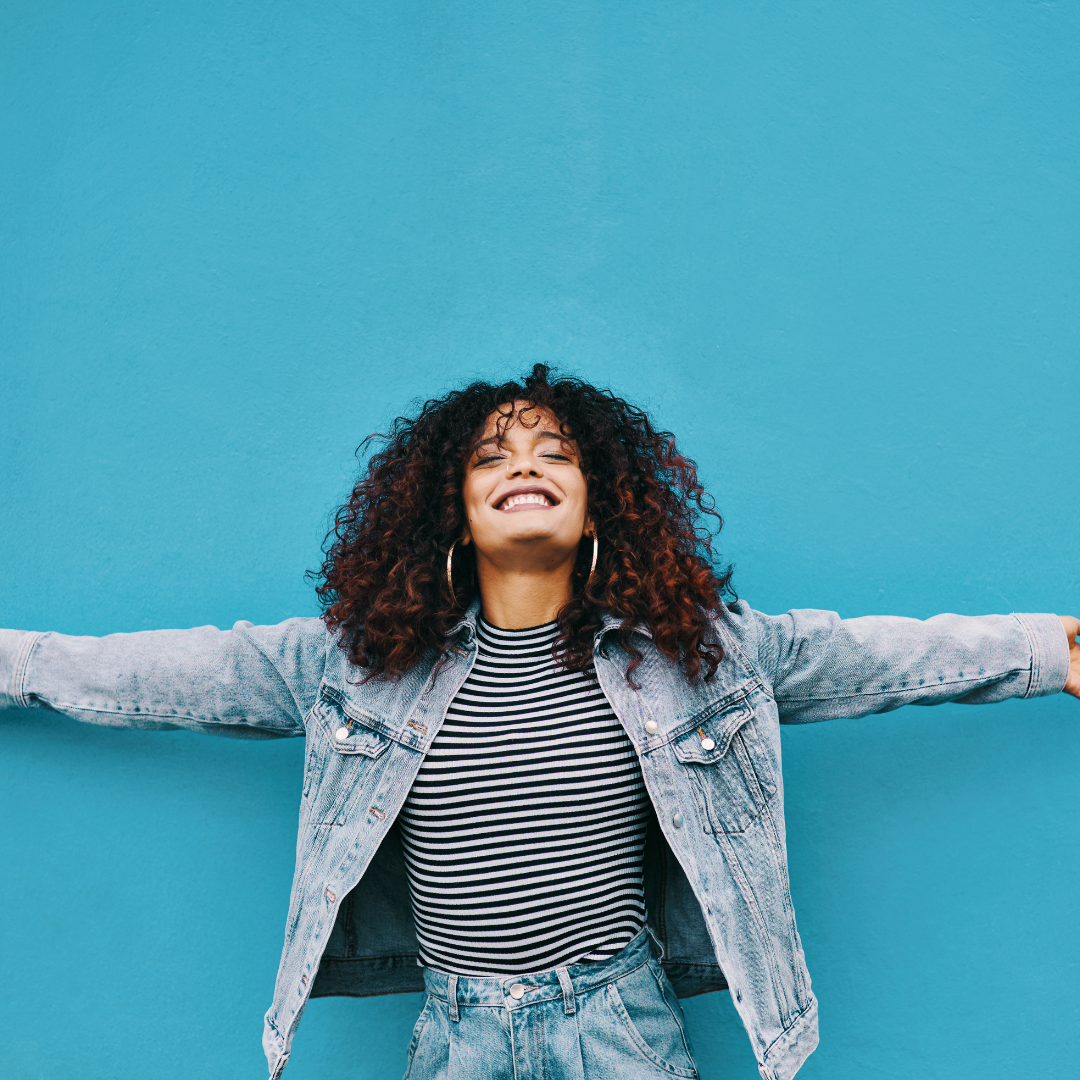 Woman with curly hair smiles, arms outstretched against a bright blue wall. She wears a striped shirt and denim jacket, exuding joy.