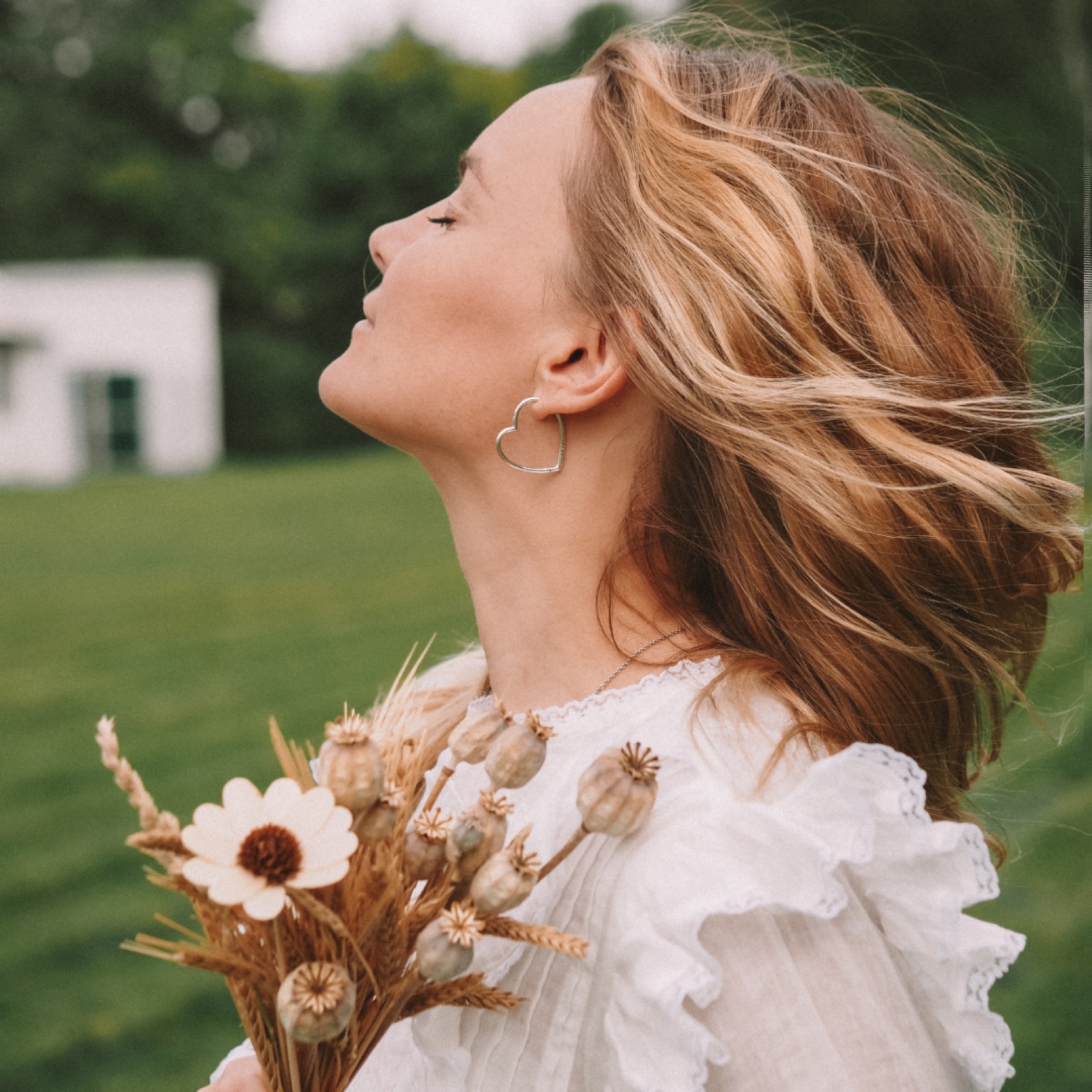 Woman in white dress with heart earrings holding dried flowers in a field. Her eyes are closed, and her hair is blowing in the wind.