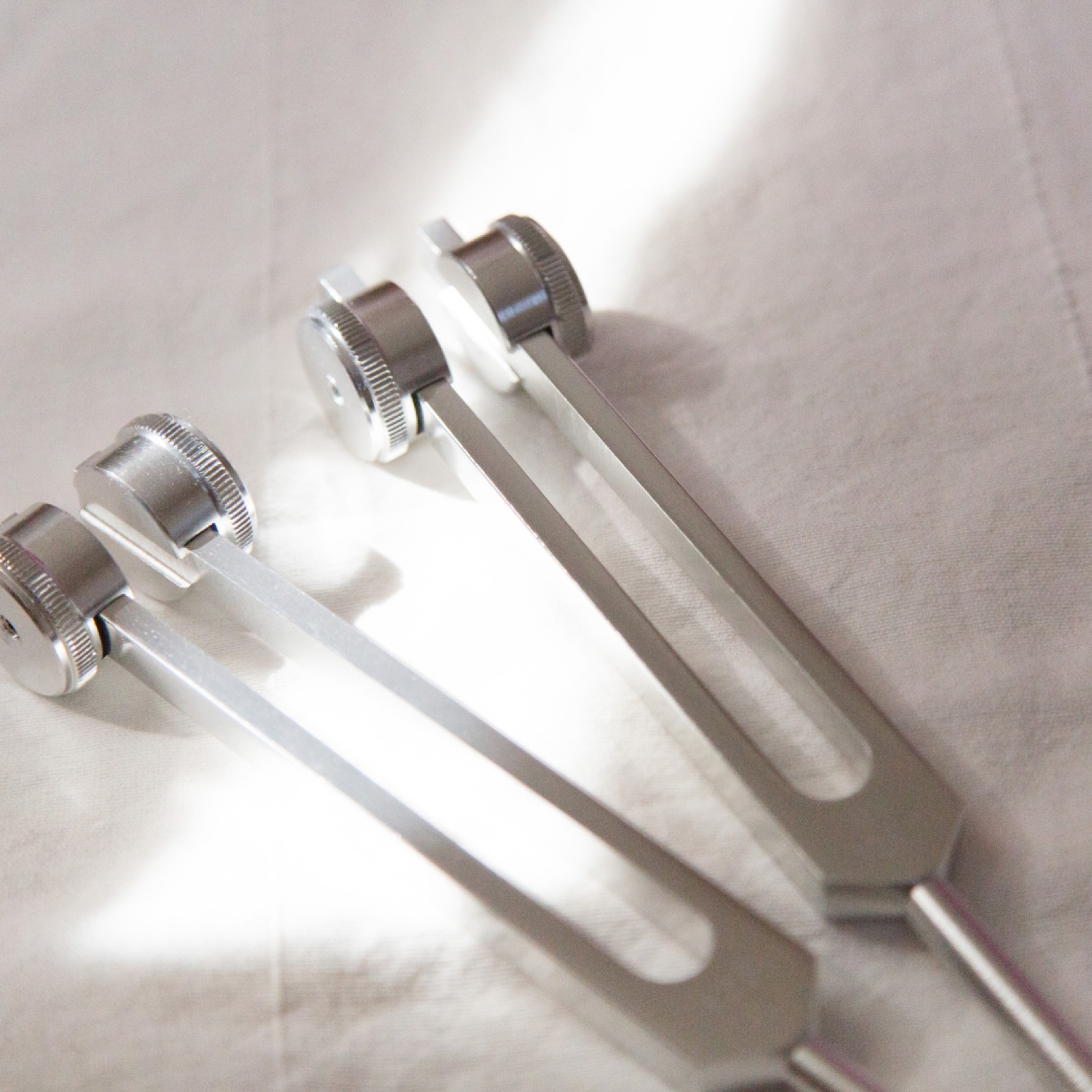 Two metal tuning forks with round tops on a white surface, bathed in bright light, casting soft shadows.