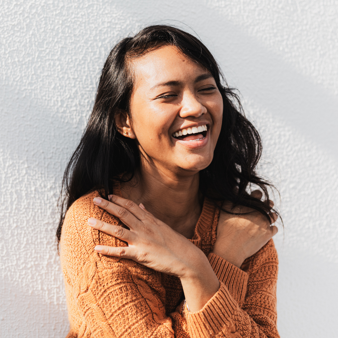 Woman in an orange sweater laughing with arms crossed on chest, against a textured white wall. Bright and joyful mood.