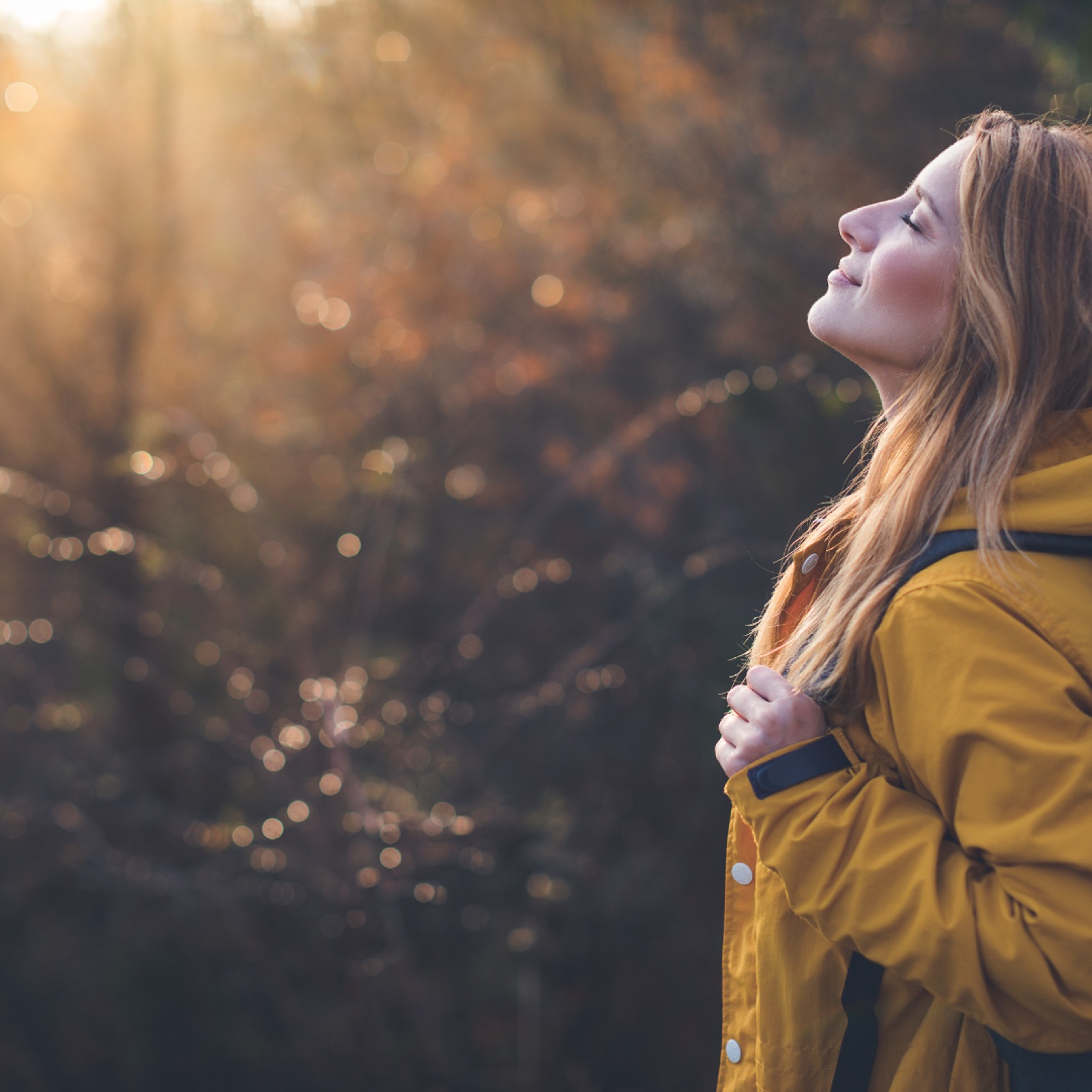 Woman in a yellow coat tilts her head back, enjoying sunlight in a blurred forest setting. Warm tones create a serene mood.