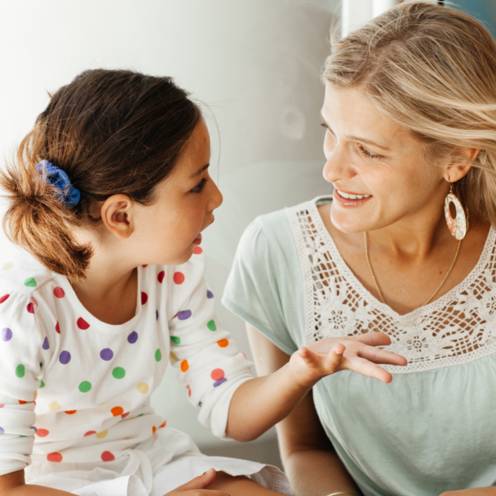 Child in a polka dot shirt talking to a smiling woman in a lace top. Bright colors and warm, engaging atmosphere.
