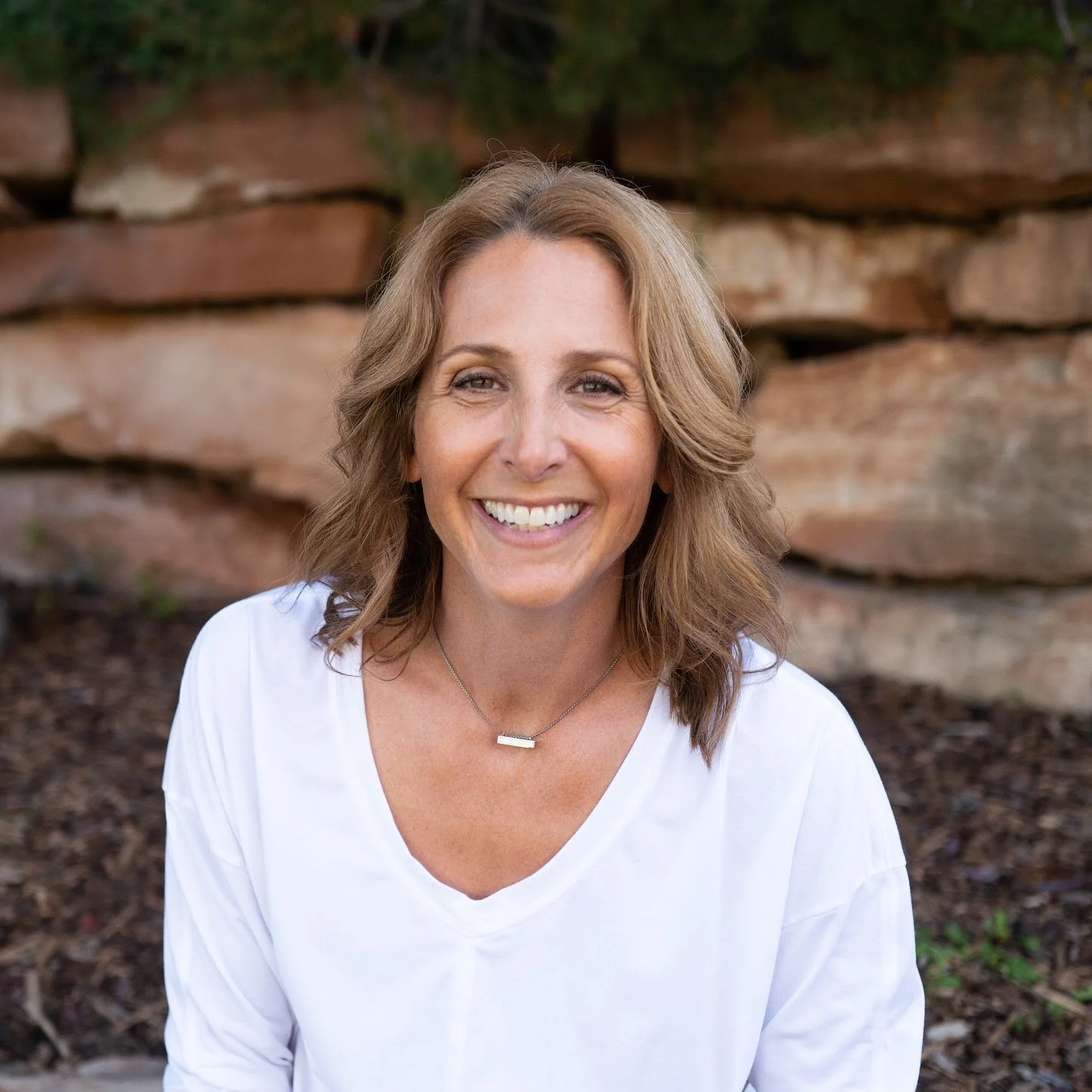 Smiling woman with light brown hair in a white top against a stone wall and greenery background. Casual and warm atmosphere.