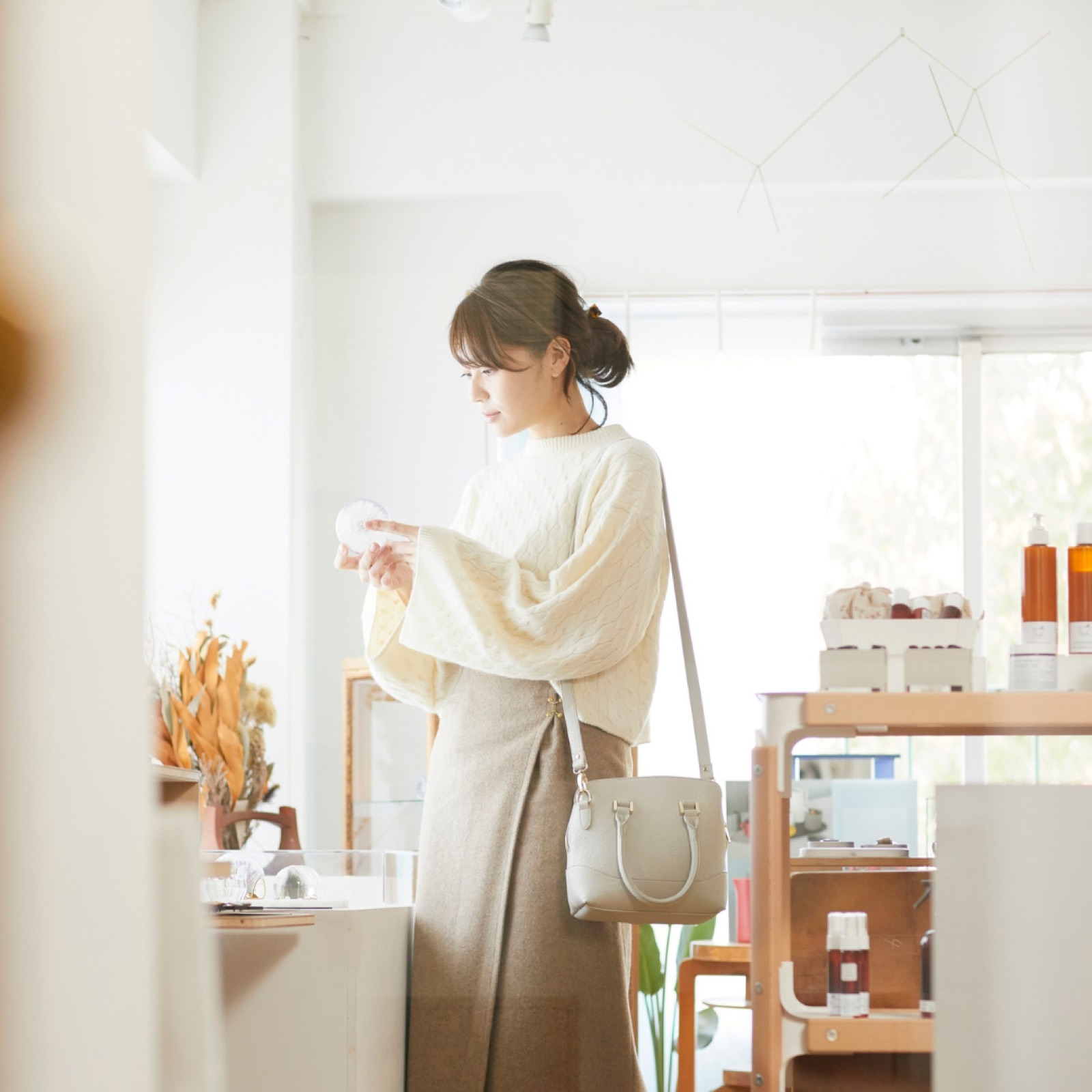 Woman in a light sweater and beige skirt browsing a shop, holding an item, with a neutral-toned interior and beauty products in the background.