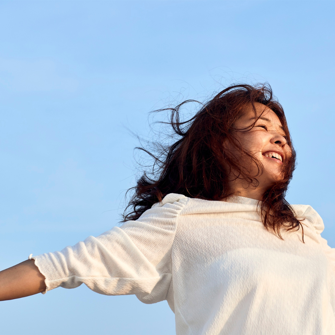 Woman smiling with outstretched arms against a clear blue sky. She's wearing a white top. The mood is joyful and carefree.