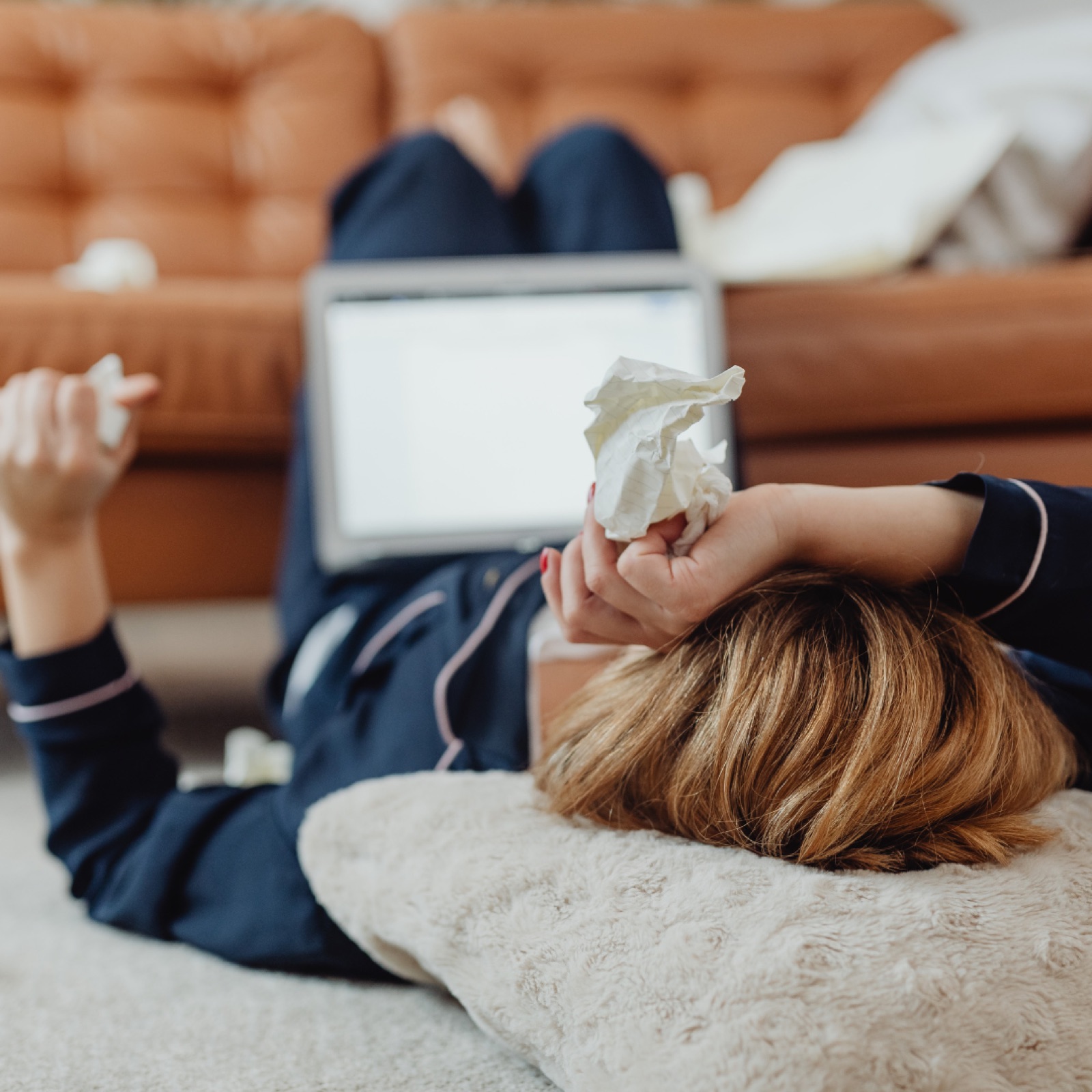 Woman in pajamas lying on floor with crumpled tissues, laptop on her stomach, in cozy room with brown sofa, conveys a relaxed mood.