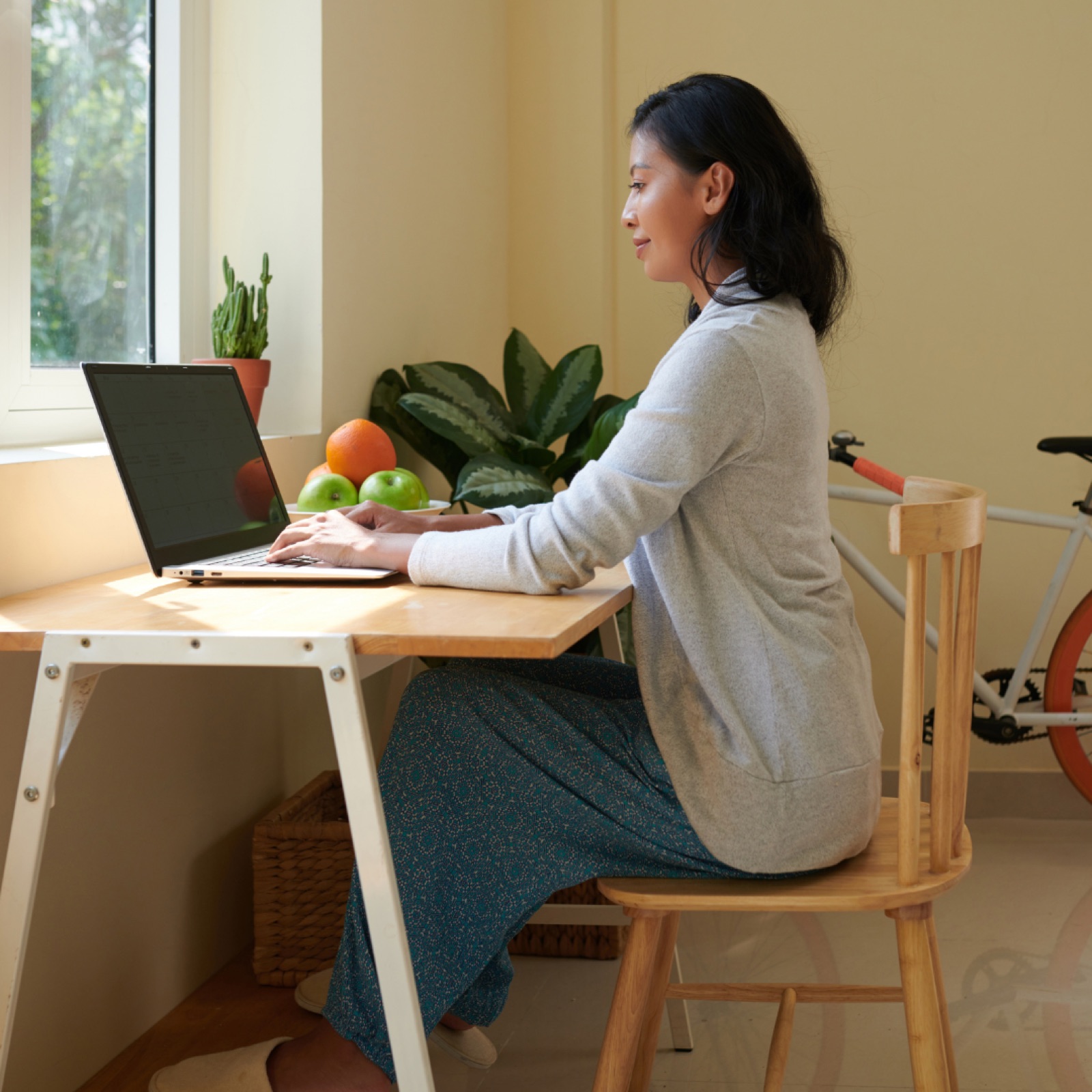 Woman working on a laptop at a wooden desk with fruit and plants on the windowsill. A bicycle is in the background, creating a calm setting.
