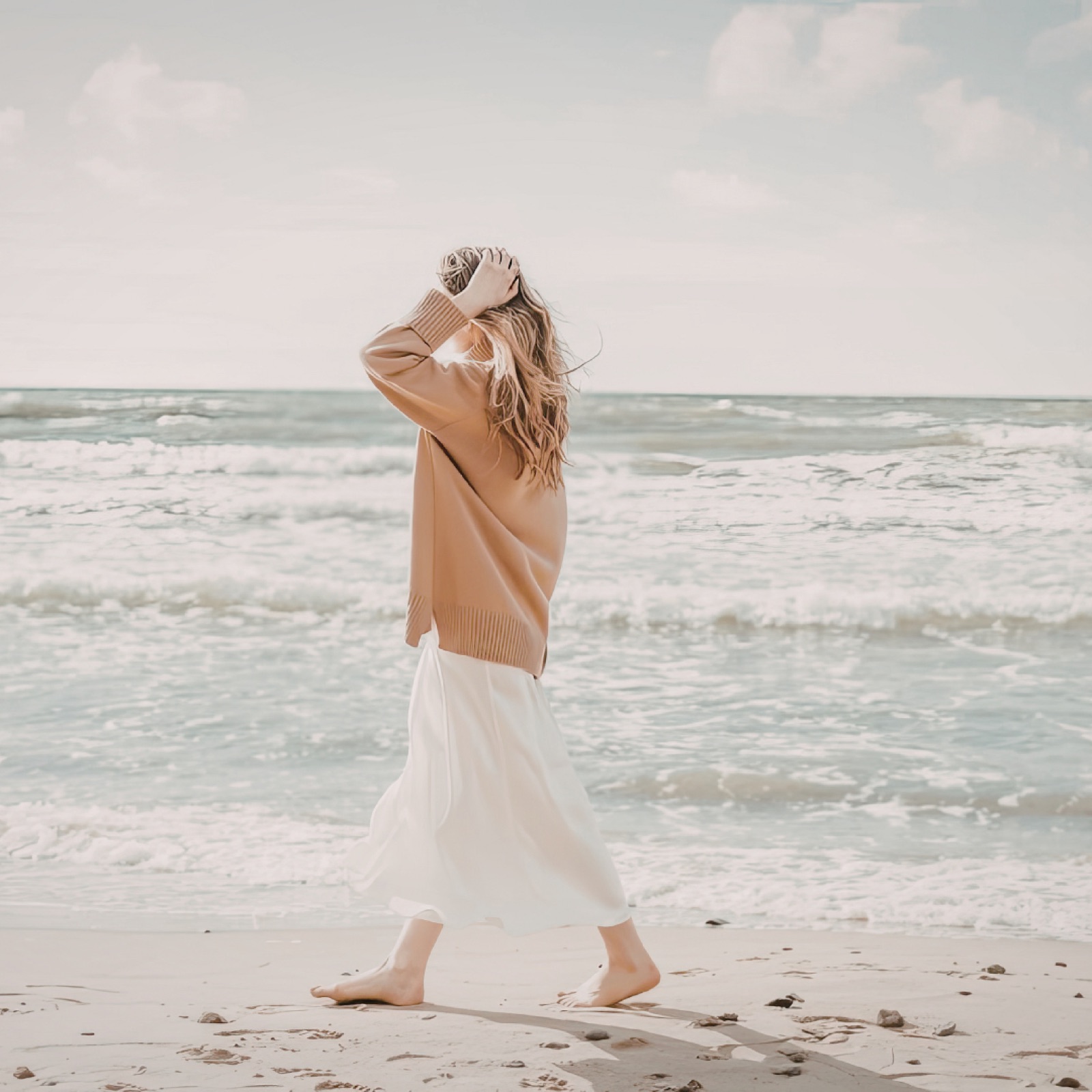 Woman in beige sweater and white skirt walks barefoot on a sandy beach. Ocean waves in the background, bright and serene atmosphere.