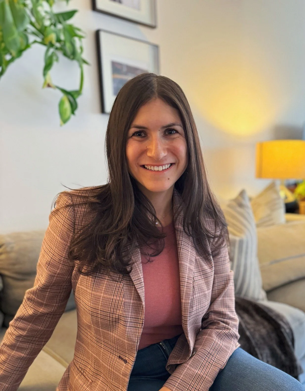 Woman smiling, seated on a sofa in a cozy living room. She's wearing a pink checkered blazer. Background has framed art and a glowing lamp.
