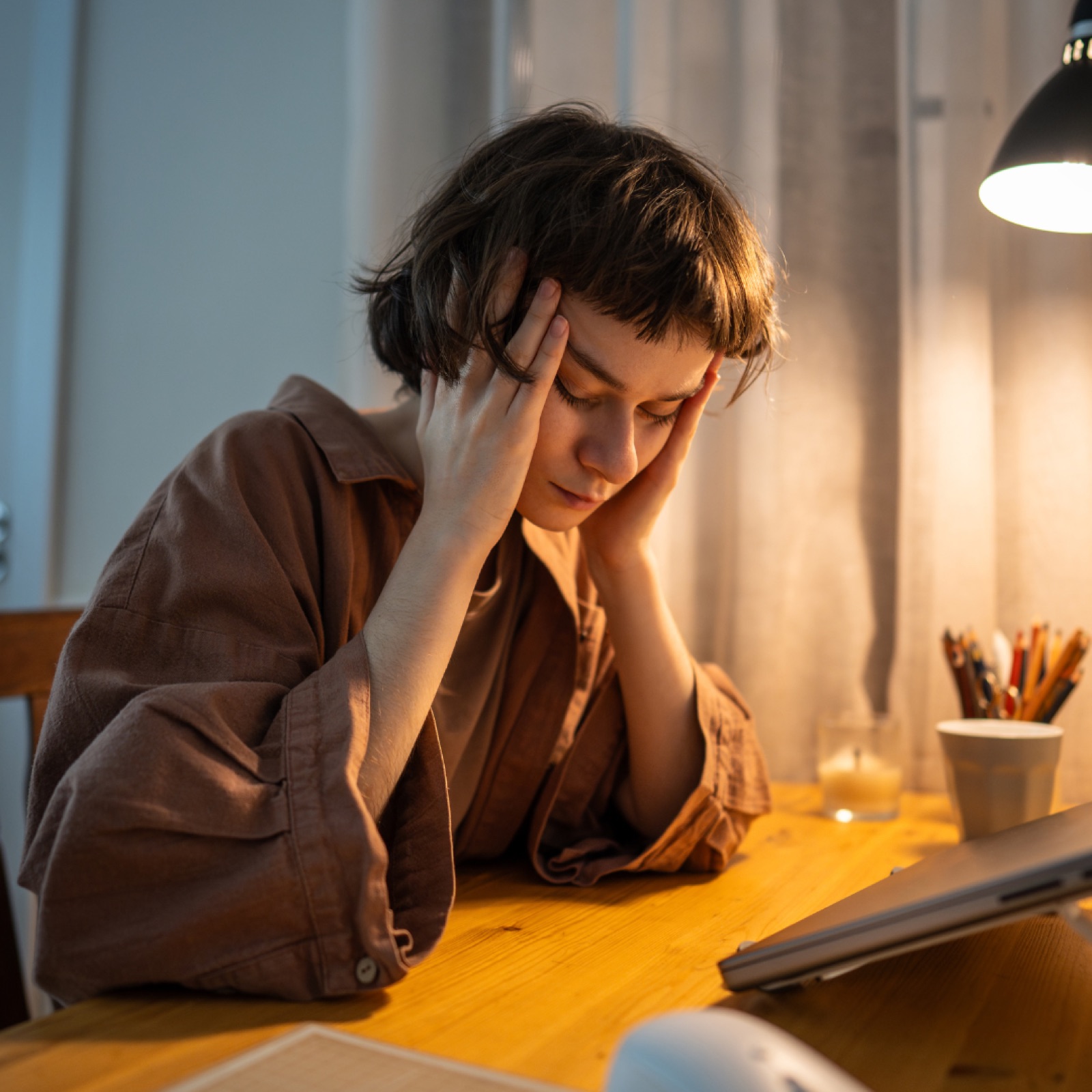 Person sitting at a wooden table, head in hands, under a lamp; appears stressed. Warm lighting, pencils in a cup, and a tablet are visible.