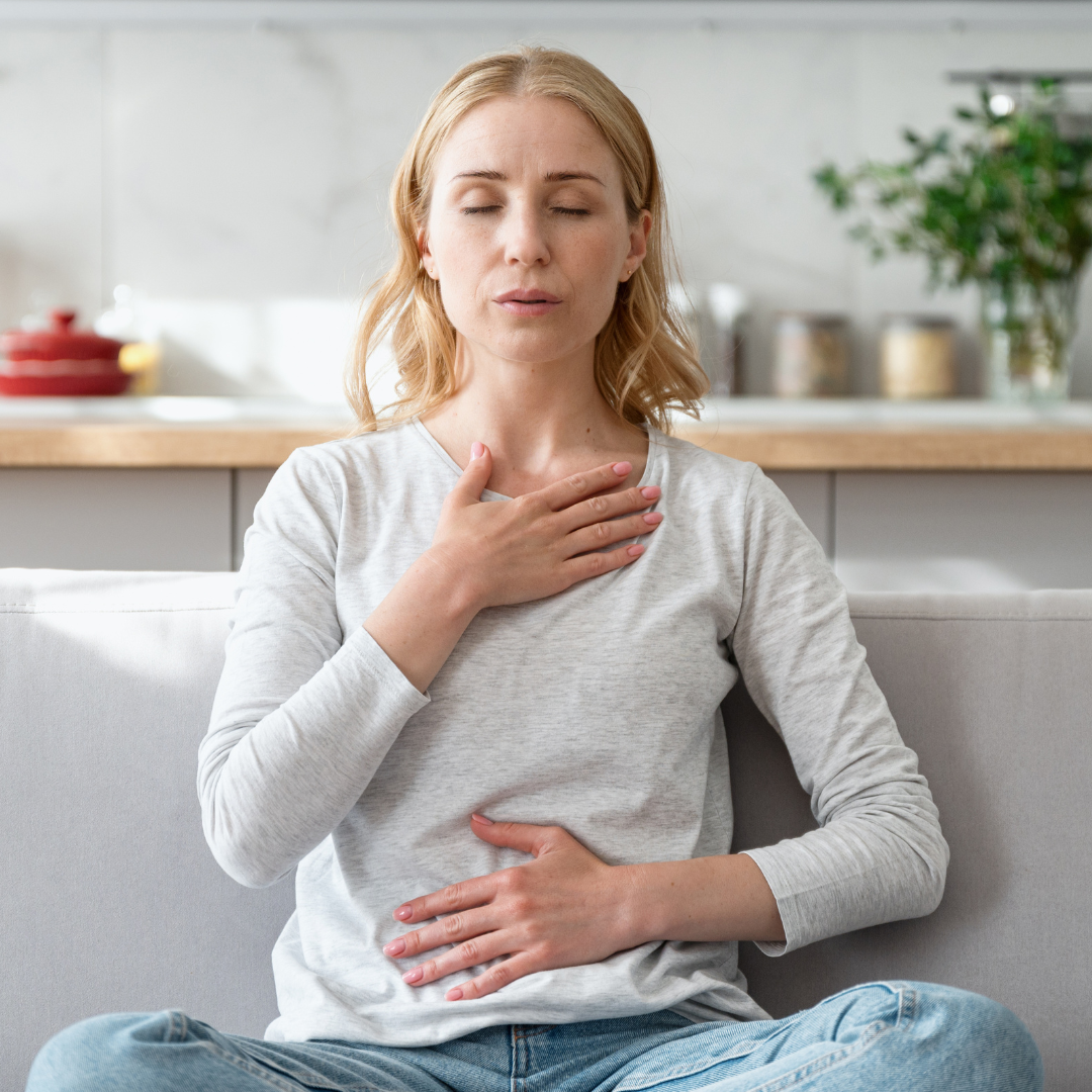 Woman meditating on a sofa with eyes closed, hand on chest, and hand on stomach. Bright background, relaxed mood.