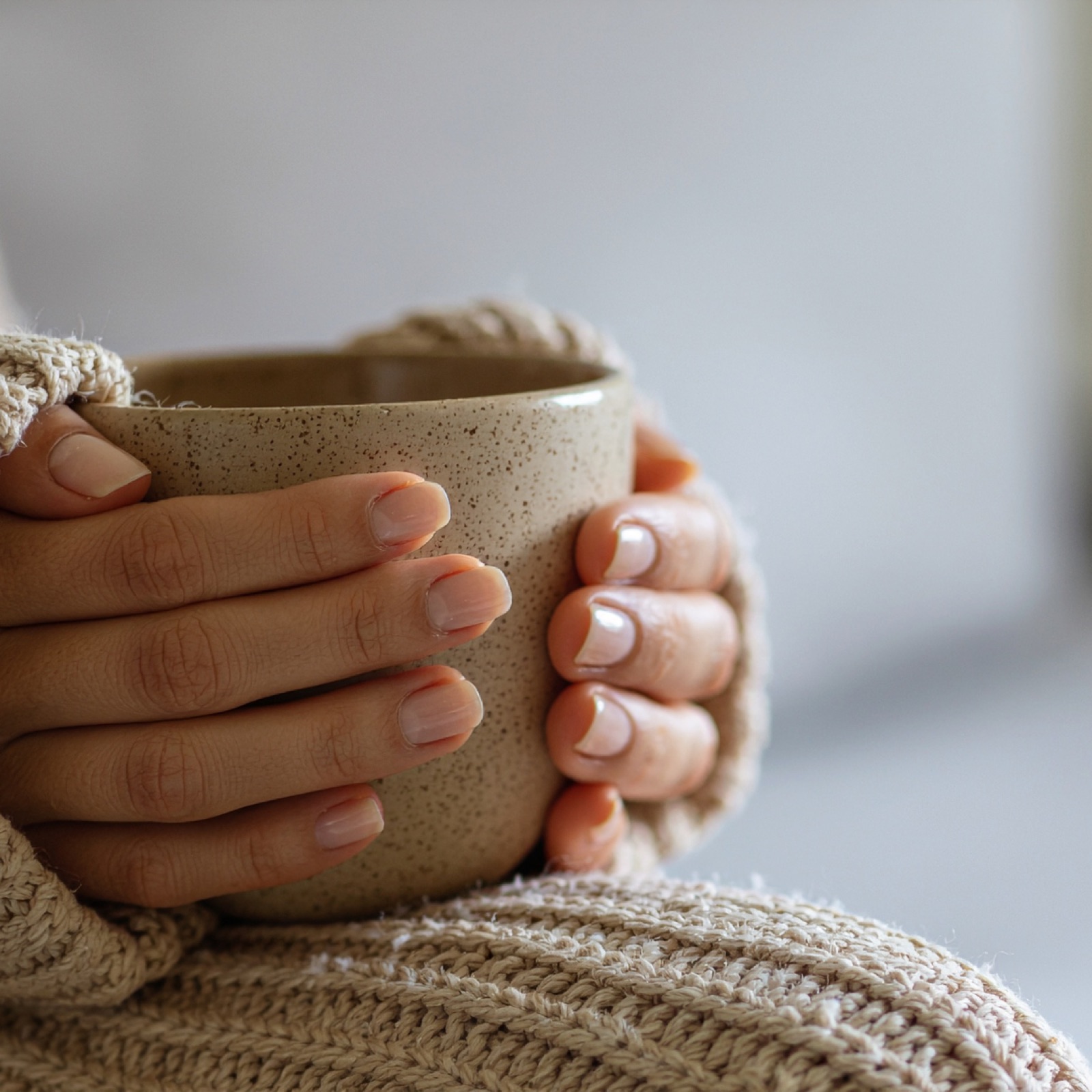 Hands holding a speckled beige mug, cozy in a knit sweater. Neutral tones and soft textures create a warm, comforting mood.