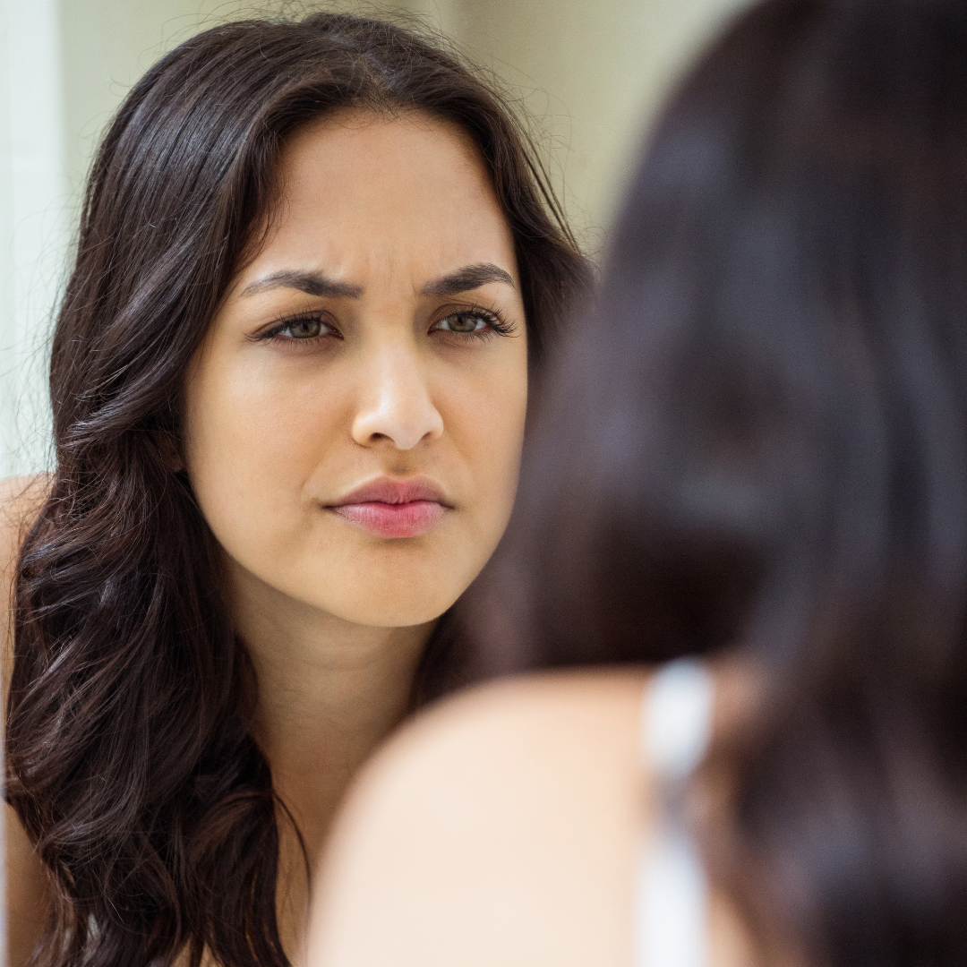Woman with dark hair, wearing a white top, looks intently into a mirror with a focused expression. Neutral background.