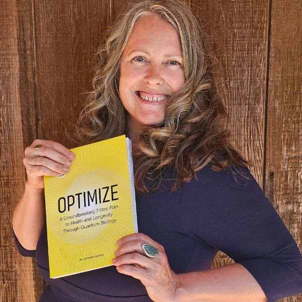 Smiling person holds a yellow book titled "OPTIMIZE" against a wooden wall. The book cover mentions health and longevity. Wearing a blue top.