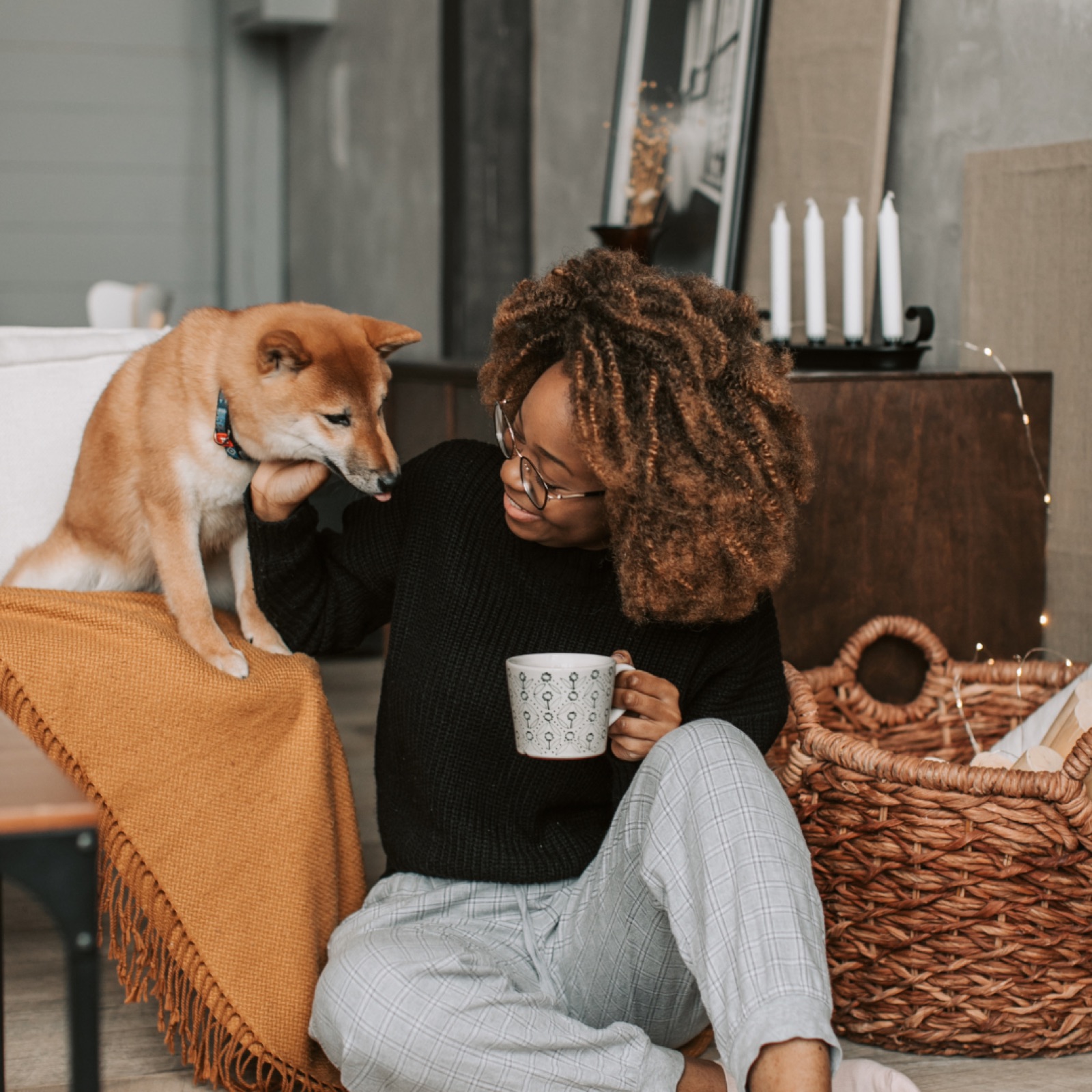 Woman with glasses smiles while petting a Shiba Inu on a couch. Holds a mug; cozy room with candles, wicker basket, and orange blanket.