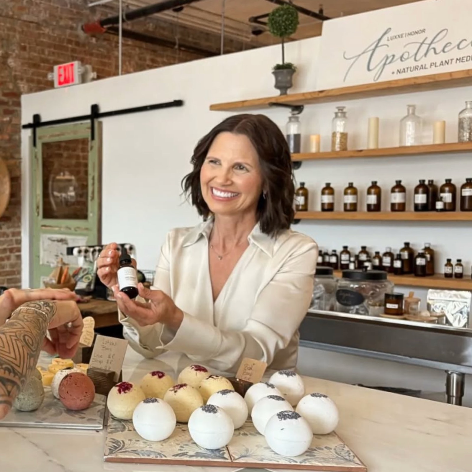 Jennifer Del Giudice, Owner
Luxxe Honor Apothecary + Wellness, holding a bottle, smiling, surrounded by bath bombs. Brick wall, shelves with jars, sign reads "Apothecary" in background.